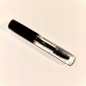 May include: A clear plastic tube with a black cap and a black mascara wand. The tube is filled with a clear liquid.