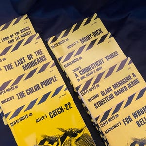 May include: A collection of Cliffs Notes study guides with yellow covers and black and white diagonal stripes. Titles include "The Lord of the Rings," "Moby Dick," and "Catch-22." The books are stacked on a dark blue surface.