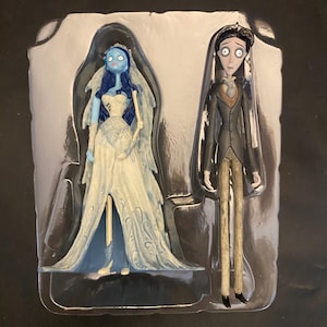 May include: A set of two figurines from the animated film, "Corpse Bride." The bride is blue-skinned, wearing a white gown with a veil. The groom is in a black suit with a brown vest and striped pants. The figures are in a plastic display case.