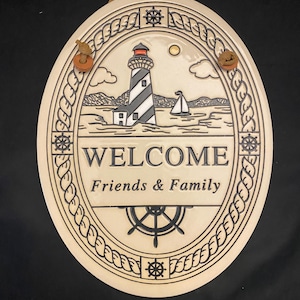 May include: Oval ceramic welcome sign with a nautical theme. The sign features a lighthouse, sailboat, and the words "WELCOME Friends & Family." The border is a rope design with ship wheels. It has a brown leather cord for hanging.