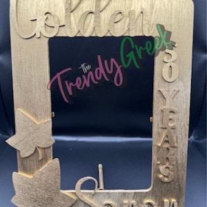 May include: A wooden picture frame with the words "Golden" and "50 Years" cut out. The frame also has the words "The Trendy Greek" and "Soror" cut out. The frame has a leaf design on the bottom left corner.