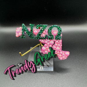 May include: A decorative item featuring the Greek letters NZΩ in green glitter and pink bubble accents. The letters are mounted on a clear stand with a gold-colored support. The words "The Trendy Greek" are also displayed.
