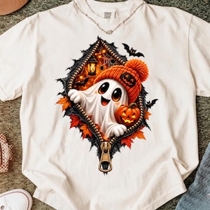 Peut inclure: Un t-shirt blanc avec un motif représentant un fantôme portant un bonnet en tricot orange, tenant une citrouille d'Halloween, sortant d'une fermeture éclair. Le design inclut des feuilles d'automne, des chauves-souris et des toiles.