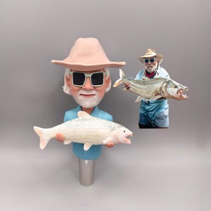 Peut inclure: Figurine d'un homme portant un chapeau et des lunettes de soleil, tenant un poisson blanc avec des accents roses. Une image plus petite montre un homme tenant un gros poisson. La figurine a une chemise bleue et une base argentée. Le poisson a la bouche ouverte.