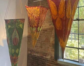 May include: Three colorful fabric cones hanging from the ceiling. The cones are made of fabric and have different patterns. The cone on the left is green with floral designs, the middle cone is red with yellow floral designs, and the cone on the right is red with orange and yellow designs.