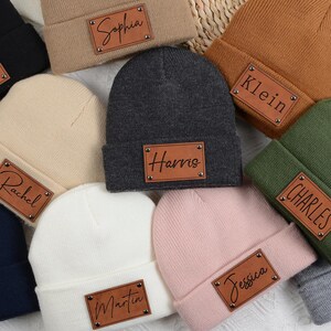 Puede incluir: Una colección de gorros personalizados en varios colores, incluyendo negro, beige, azul marino, blanco, gris, marrón, verde y rosa. Cada gorro presenta un parche rectangular de cuero con un nombre grabado en cursiva.