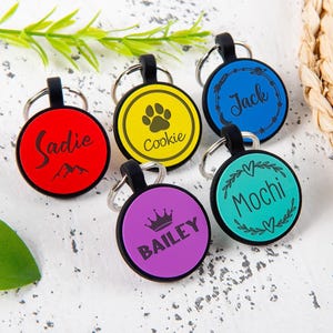 Puede incluir: Cinco placas de identificaci&oacute;n circulares para mascotas con bordes negros y anillas met&aacute;licas. Las placas son roja con el nombre Sadie, amarilla con una huella y Cookie, azul con iconos espaciales y Jack, morada con una corona y BAILEY, y turquesa con Mochi.