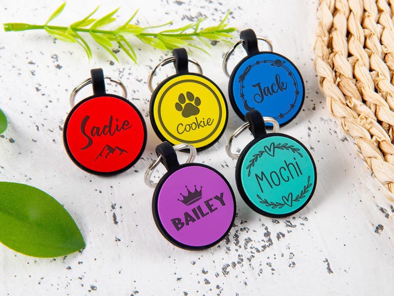 Puede incluir: Cinco placas de identificaci&oacute;n circulares para mascotas con bordes negros y anillas met&aacute;licas. Las placas son roja con el nombre Sadie, amarilla con una huella y Cookie, azul con iconos espaciales y Jack, morada con una corona y BAILEY, y turquesa con Mochi.
