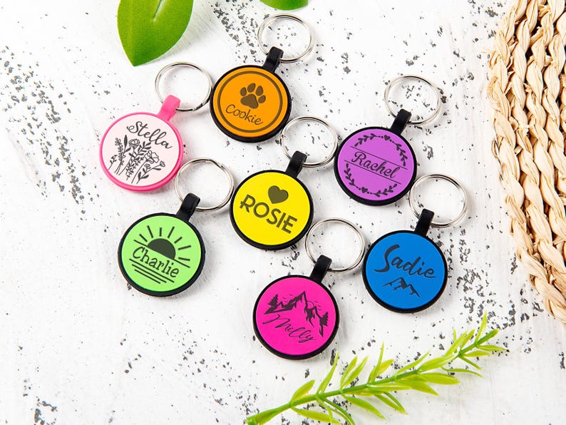 Puede incluir: Una colecci&oacute;n de siete llaveros circulares en varios colores, incluyendo rosa, naranja, amarillo, morado, verde, azul y rosa intenso. Cada llavero cuenta con una anilla met&aacute;lica y un dise&ntilde;o gr&aacute;fico negro &uacute;nico con nombres como Stella, Cookie, Rosie, Rachel, Charlie, Milly y Sadie.