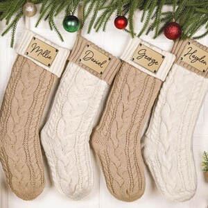 Peut inclure: Quatre chaussettes de Noël en tricot torsadé aux couleurs alternées beige et crème. Chaque chaussette présente un revers côtelé avec une étiquette nominative en bois rectangulaire gravée des prénoms Millie, Daniel, George et Hayden.