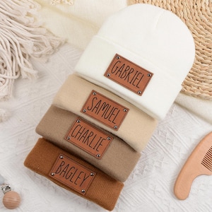 Könnte beinhalten: Vier Strickmützen in Creme, Beige, Braun und Rost. Jede Mütze hat ein braunes Lederetikett mit einem Namen: Gabriel, Samuel, Charlie und Bagley. Ein Holzkamm und ein Schnullerclip sind ebenfalls zu sehen.