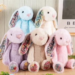 Peut inclure: Cinq lapins en peluche aux couleurs pastel : bleu clair, blanc, lavande, crème et rose. Chaque lapin a une doublure à motifs floraux dans les oreilles et les pieds, et un nom brodé sur une oreille. Les lapins sont posés sur une surface en bois.