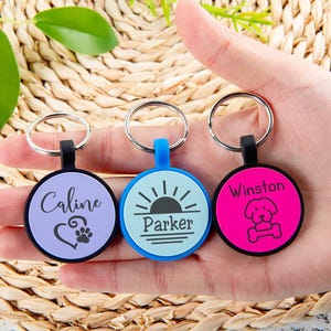 Puede incluir: Tres placas de identificaci&oacute;n circulares para mascotas con anillas met&aacute;licas. La placa izquierda es de color morado claro con el nombre Caline, un coraz&oacute;n y una huella. La central es azul claro con el nombre Parker y un sol. La derecha es rosa intenso con el nombre Winston y un icono de perro y hueso.