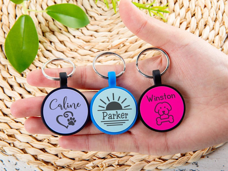 Puede incluir: Tres placas de identificaci&oacute;n circulares para mascotas con anillas met&aacute;licas. La placa izquierda es de color morado claro con el nombre Caline, un coraz&oacute;n y una huella. La central es azul claro con el nombre Parker y un sol. La derecha es rosa intenso con el nombre Winston y un icono de perro y hueso.