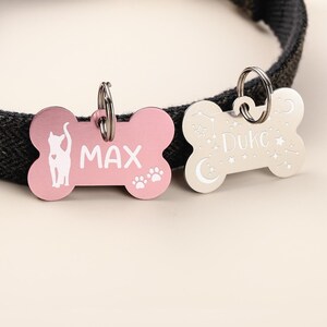 May include: Two bone-shaped pet ID tags attached to a black collar. The pink tag features a white cat silhouette, the name MAX, and paw prints. The silver tag features the name Duke with engraved stars, a crescent moon, and constellation patterns.