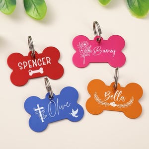 May include: Four bone-shaped pet ID tags in red, pink, blue, and orange. The red tag says SPENCER with a bone icon. The pink tag says Barney with a floral graphic. The blue tag says Olive with a cross and dove. The orange tag says Bella with a leaf wreath.
