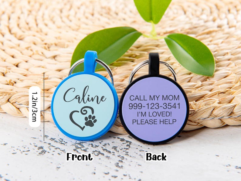 Puede incluir: Dos placas de identificaci&oacute;n circulares para mascotas de 3 cm de di&aacute;metro. La azul muestra el nombre Caline y un coraz&oacute;n con una huella. La negra incluye el texto CALL MY MOM, 999-123-3541, I'M LOVED!, PLEASE HELP. Ambas tienen anillas met&aacute;licas.