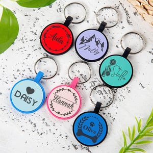 Puede incluir: Una colecci&oacute;n de siete llaveros circulares en varios colores, incluyendo rojo, morado claro, verde azulado, azul claro, rosa y azul oscuro. Cada llavero cuenta con una anilla met&aacute;lica y un dise&ntilde;o negro impreso con nombres como Aida, Milly, Stella, Daisy, Hannah y Olive.