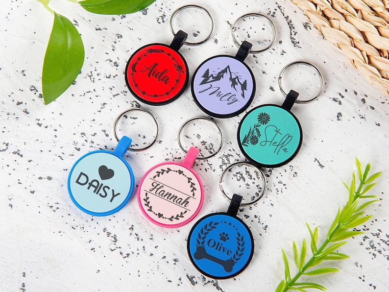 Puede incluir: Una colecci&oacute;n de siete llaveros circulares en varios colores, incluyendo rojo, morado claro, verde azulado, azul claro, rosa y azul oscuro. Cada llavero cuenta con una anilla met&aacute;lica y un dise&ntilde;o negro impreso con nombres como Aida, Milly, Stella, Daisy, Hannah y Olive.