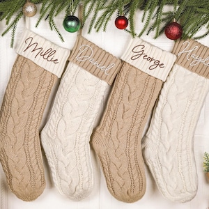 May include: Four cable-knit Christmas stockings in alternating beige and cream colours. Each stocking features a ribbed cuff with a personalised name embroidered in brown or white script: Millie, Daniel, George, and Hayden, respectively.