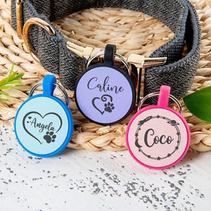 Puede incluir: Tres placas de identificaci&oacute;n circulares para mascotas con bordes de silicona en azul claro, lavanda y rosa. La placa azul claro tiene el nombre Angela con un coraz&oacute;n y una huella. La placa lavanda muestra Caline con un coraz&oacute;n y una huella. La placa rosa muestra Coco dentro de un borde de estrellas y planetas.