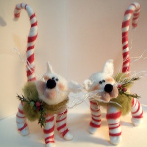May include: Two white felt cat figurines with candy cane tails and green and red striped legs. Each cat is wearing a green felt vest with a sprig of greenery and red berries.