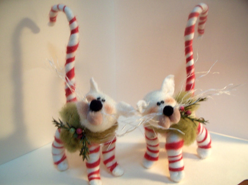 May include: Two white felt cat figurines with candy cane tails and green and red striped legs. Each cat is wearing a green felt vest with a sprig of greenery and red berries.
