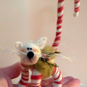 May include: A white felt cat with a red and white candy cane tail, wearing a green felt vest with a sprig of holly. The cat is standing on a hand.