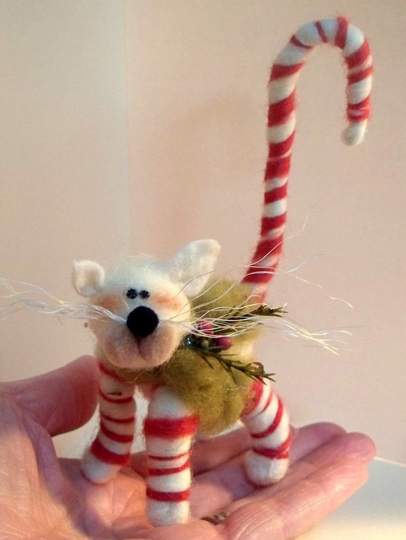 May include: A white felt cat with a red and white candy cane tail, wearing a green felt vest with a sprig of holly. The cat is standing on a hand.