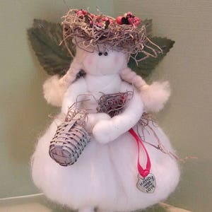 May include: A white felt angel doll with a brown nest hat, holding a small woven basket. The doll has a pink face and red cheeks. The doll is wearing a pink ribbon with a silver heart charm that says "Hand Made With Love".