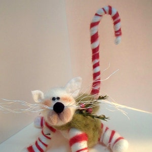 May include: White felt cat with a candy cane tail, wearing a green and white striped outfit and a green felt wreath around its neck.