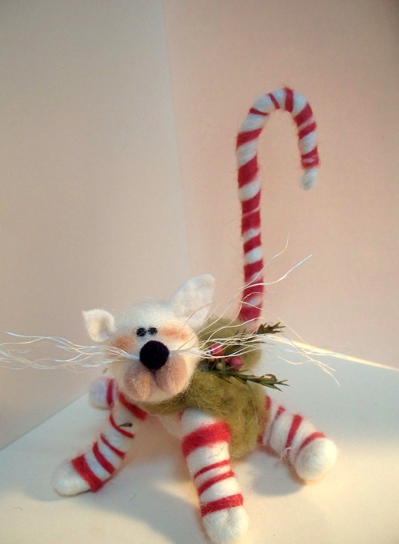 May include: White felt cat with a candy cane tail, wearing a green and white striped outfit and a green felt wreath around its neck.