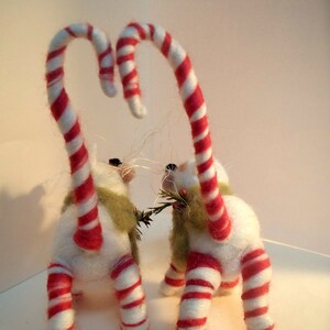 May include: Two white felted cat ornaments with red and white candy cane tails. The cats are holding a sprig of green leaves in their paws.