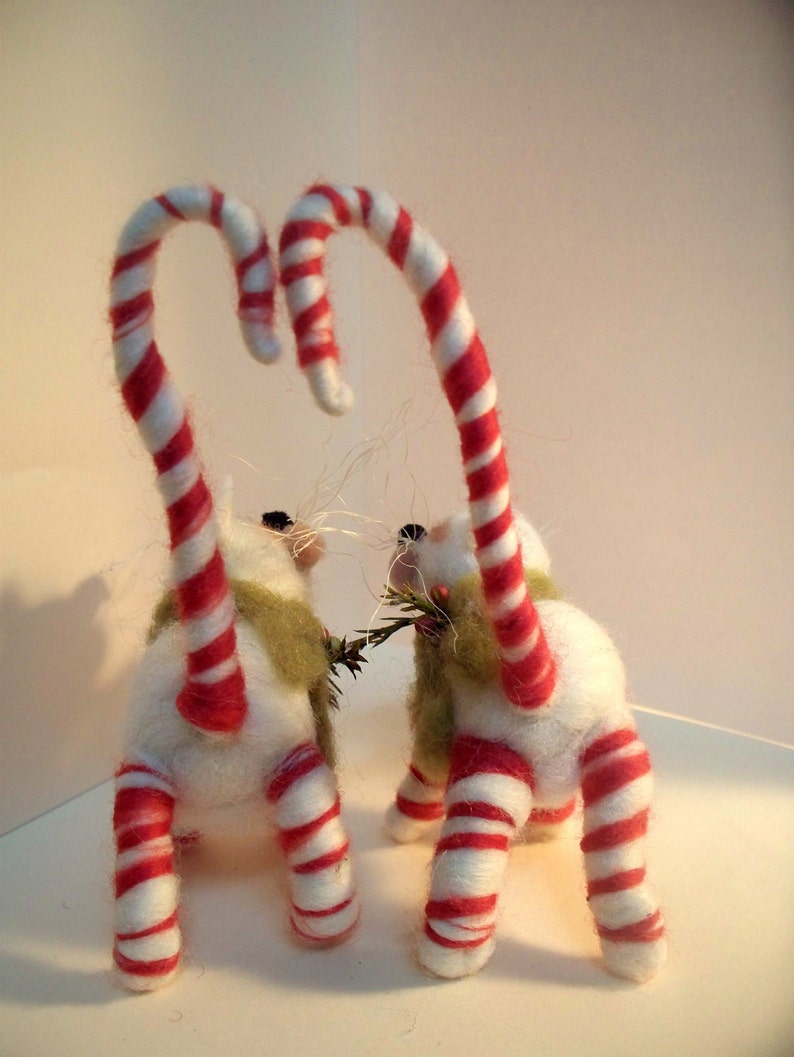 May include: Two white felted cat ornaments with red and white candy cane tails. The cats are holding a sprig of green leaves in their paws.