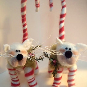 May include: Two white felt cat figurines with red and white candy cane legs and tails. The cats have black eyes and noses and are wearing green felt sweaters with sprigs of greenery.