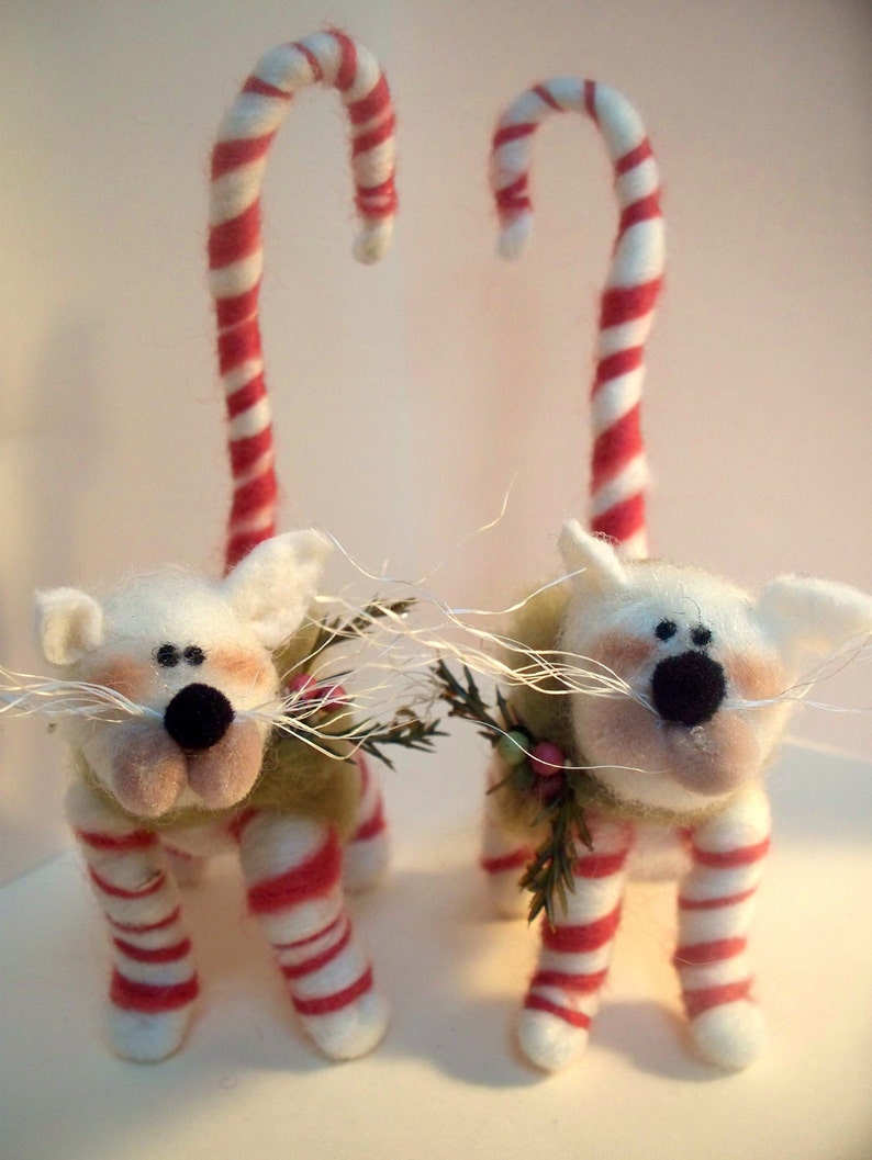May include: Two white felt cat figurines with red and white candy cane legs and tails. The cats have black eyes and noses and are wearing green felt sweaters with sprigs of greenery.