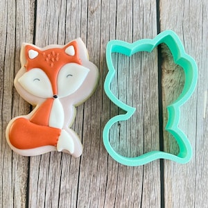 May include: A light green plastic cookie cutter shaped like a fox, next to a decorated sugar cookie in the shape of a fox. The cookie is decorated with orange, white, and brown icing.