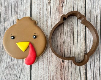 May include: A brown cookie cutter shaped like a turkey head with a red wattle and a yellow beak. The cookie cutter is next to a decorated cookie with brown icing, black eyes, a yellow beak, and a red wattle.