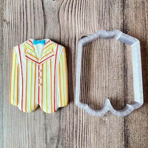May include: A colorful cookie decorated to resemble a striped jacket with a blue bow tie. Next to it is a plastic cookie cutter in the shape of the jacket. The jacket has vertical stripes in red, orange, and yellow.