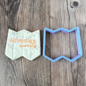 May include: A map-shaped cookie with the words "Adventure awaits" in orange script, next to a blue map-shaped cookie cutter. The cookie has a world map design in gray.