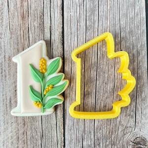 May include: A decorated cookie in the shape of the number one, adorned with green leaves and yellow berries. Next to it is a yellow plastic cookie cutter in the shape of the number one. The cookie is on a wooden surface.