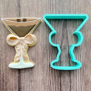 May include: A turquoise plastic cookie cutter shaped like a martini glass. Next to it is a decorated sugar cookie in the shape of a martini glass with a pink bow.