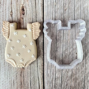 May include: A beige onesie cookie decorated with white flower accents and ruffled sleeves. Next to it is a clear plastic cookie cutter in the shape of the onesie. The cookie and cutter are on a wooden surface.