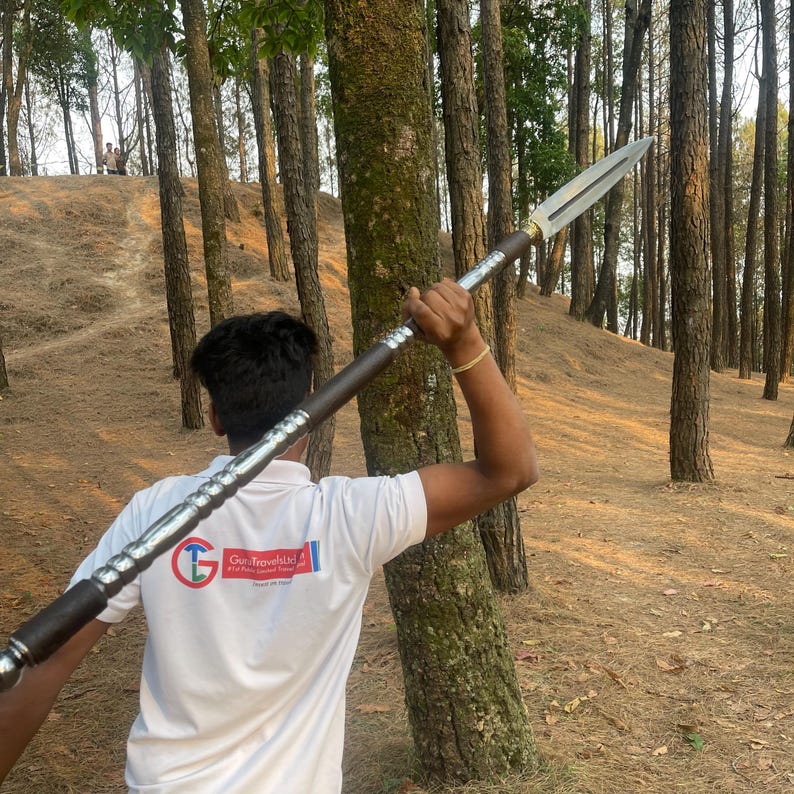 May include: A person in a white shirt with the text "Go-Travel-Kid" holds a long, silver-colored spear with a brown handle. The spear is pointed towards the right side of the image. The person is standing in a forest setting.