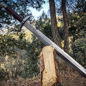 Puede incluir: Una espada plateada con un mango de madera oscura está apoyada en un tronco de madera. La espada tiene una guarda curva y una punta afilada. El fondo es una imagen borrosa de árboles.
