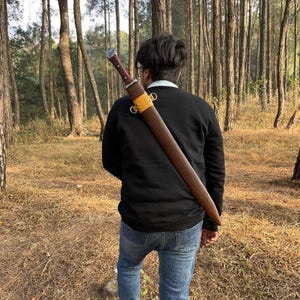 May include: A brown sword in a sheath is carried over the shoulder. The sword has a silver hilt and a dark brown handle. The sheath is brown with a yellow strap. The person is wearing a black sweater and blue jeans.