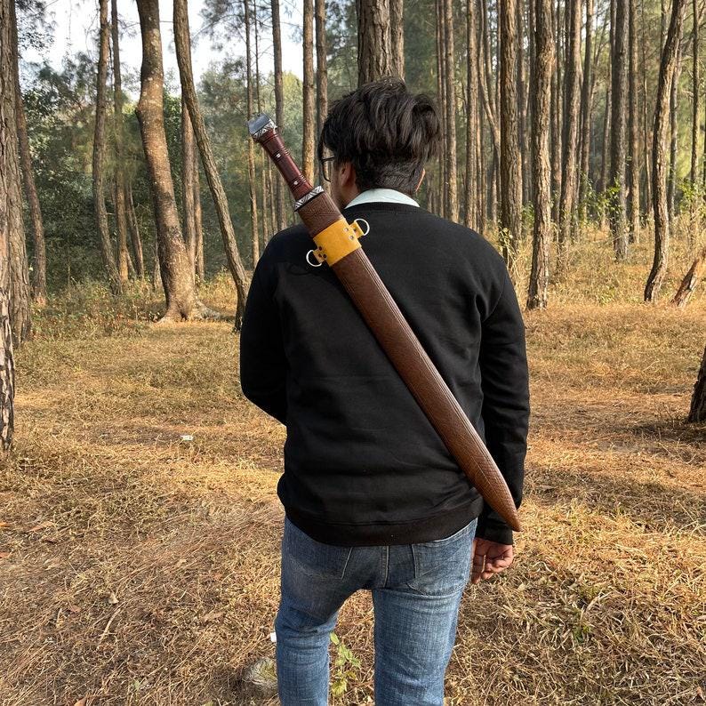 May include: A brown sword in a sheath is carried over the shoulder. The sword has a silver hilt and a dark brown handle. The sheath is brown with a yellow strap. The person is wearing a black sweater and blue jeans.