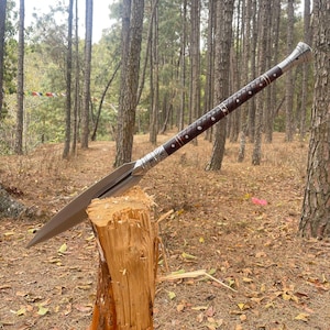 May include: A silver and brown spear with a wooden handle is stuck into a tree stump in a forest setting. The spear has a decorative design on the handle and blade.