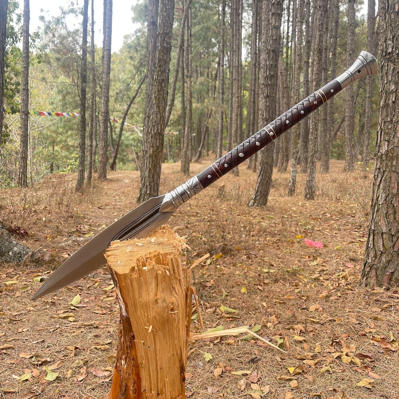 May include: A silver and brown spear with a wooden handle is stuck into a tree stump in a forest setting. The spear has a decorative design on the handle and blade.