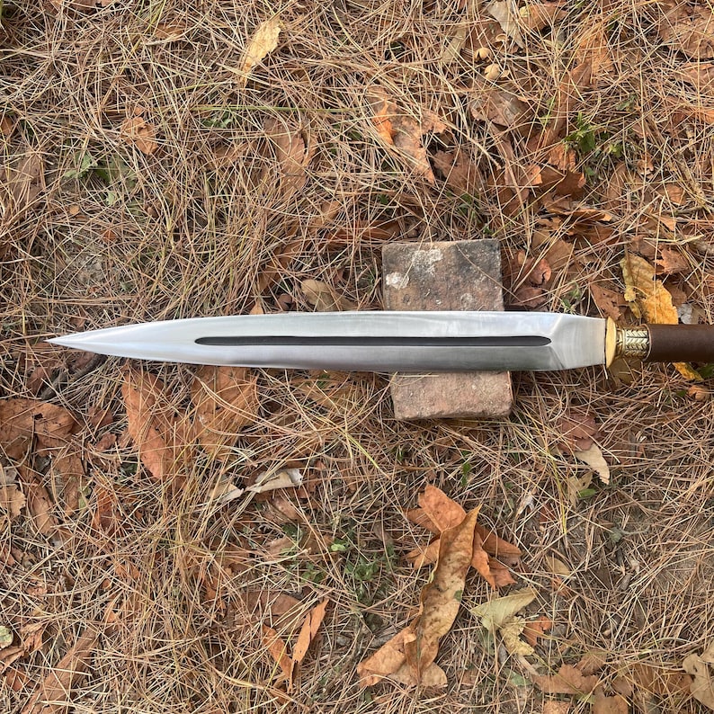 May include: A long, silver-colored sword with a brown handle and gold accents. The sword is lying on a bed of brown leaves.